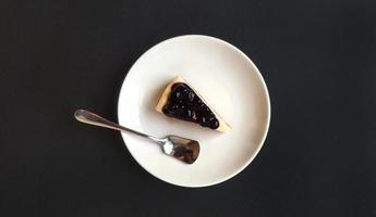 Blick von oben auf die Scheibe Blaubeerkäsekuchen und Edelstahllöffel in weißer Schale oder Platte auf schwarzem Hintergrund mit Kopierraum. flache Lage von süßem Dessert und hausgemachtem Essen auf dunklem Tisch foto