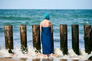 junge blauhaarige frau in langem dunkelblauem kleid, die am sandstrand steht und den ozeanhorizont betrachtet foto