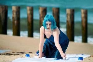 Künstlerische blauhaarige Performance-Künstlerin mit Gouache-Farben auf großer Leinwand am Strand beschmiert foto