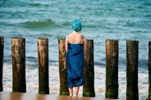 Junge blauhaarige Frau in langem dunkelblauem Kleid steht am Sandstrand und blickt auf den Meereshorizont foto