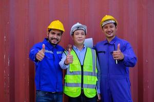 porträt von drei asiatischen industriearbeitern mit sicherheitsuniform und schutzhelmen auf blechhintergrund, daumen hoch, blick in die kamera und lächeln, frachtversand, professionelles frachttransportgeschäft. foto