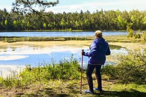 Ältere blonde kaukasische ältere Frau steht mit roten nordischen Stöcken und genießt die grüne Natur des Seewaldes foto