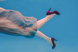 surreales kunstporträt einer jungen frau in grauem kleid, perlenschal, kleiner tasche, violetten high heels unter wasser im schwimmbad foto