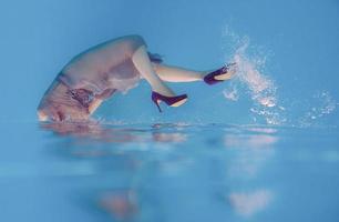 surreales kunstporträt einer jungen frau in grauem kleid, perlenschal, kleiner tasche, violetten high heels unter wasser im schwimmbad foto