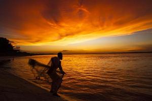 Ein Fischer, der bei Sonnenuntergang Fische auf dem Meer gegen den orangefarbenen Himmel fängt foto