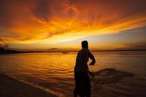 Ein Fischer, der bei Sonnenuntergang Fische auf dem Meer gegen den orangefarbenen Himmel fängt foto