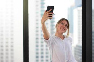 Porträt einer jungen attraktiven Frau, die Selfie-Foto auf dem Smartphone im Hintergrund eines modernen Bürogebäudes macht foto
