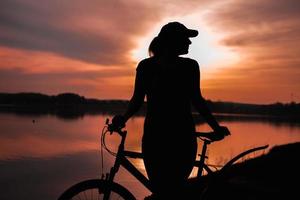 sommerliche Silhouettenlandschaft. Mädchen mit Fahrrad am See vor dem Hintergrund der untergehenden Sonne, des purpurroten Sonnenuntergangs foto