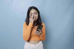 ein Porträt einer traurigen asiatischen Frau, die ihr Smartphone auf blauem Hintergrund betrachtet foto