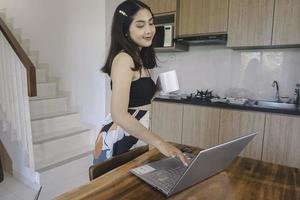 junge asiatische frau verbringt ihre zeit zu hause und arbeitet remote im esszimmer mit einer online-konferenz auf ihrem laptop, während sie einen becher trägt. foto