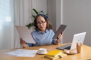 Porträt einer älteren Frau, die im Haus arbeitet und mit Laptops und Tablets arbeitet und wichtige Dokumente für Kunden überprüft. foto