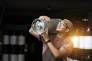 junger mann in lederschürze, der bierfass in der modernen brauerei hält, handwerksbrauereiarbeiter foto