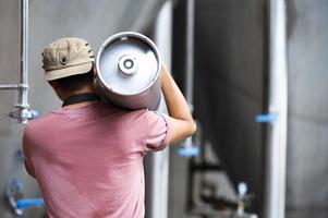 junger mann in lederschürze, der bierfass in der modernen brauerei hält, handwerksbrauereiarbeiter foto