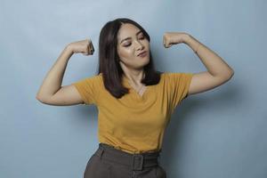 Junges schönes Mädchen mit lässigem gelbem T-Shirt, das über isoliertem blauem Hintergrund steht und stolz lächelnde Armmuskeln zeigt. Fitness-Konzept. foto