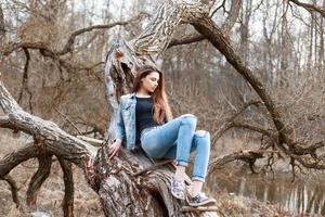 schönes Mädchen in Jeansjacke und Jeansshorts, das auf einem Baum sitzt. Entspannen Sie sich in der Natur. foto