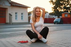 Schönes Hipster-Mädchen, das bei Sonnenuntergang auf dem Asphalt in der Nähe einer Straße sitzt foto
