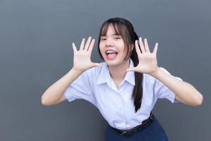 Schönes asiatisches Gymnasiastmädchen in der Schuluniform mit einem selbstbewussten Lächeln, während sie glücklich mit Grau im Hintergrund in die Kamera schaut. foto