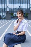 Schönes asiatisches Gymnasiastmädchen in der Schuluniform mit einem selbstbewussten Lächeln, während sie sitzt und glücklich mit dem Gebäude im Hintergrund in die Kamera schaut. foto