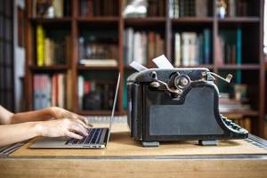 Frauenhände tippen auf modernes Notizbuch. Alte, staubbedeckte Vintage-Schreibmaschine mit weißem Blatt Papier in der Nähe eines modernen Notizbuchs auf Bücherregalhintergrund. Moderne Technik und Vintage-Geräte foto