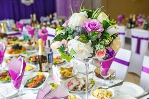 Schöne Blumen auf einem eleganten Esstisch am Hochzeitstag. Dekorationen, die auf dem festlich gedeckten Tisch in violettem Hintergrund serviert werden foto