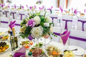 Schöne Blumen auf einem eleganten Esstisch am Hochzeitstag. Dekorationen, die auf dem festlich gedeckten Tisch in violettem Hintergrund serviert werden foto