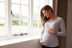 schwangere Frau, die durch Fenster steht, das Bauch hält foto