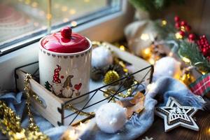 Weihnachtsdekoration am Fenster auf einer gemütlichen Holzfensterbank mit einem Becher mit einem Getränk und einer Keksdose. Lichterketten, Weihnachtsbaumzweig, Frühstückstablett-Stillleben foto