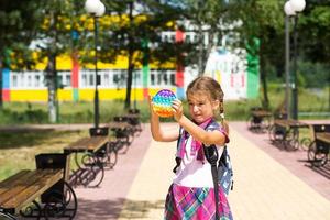 Kleines Mädchen mit Rucksack und in Schuluniform auf dem Schulhof spielt Pop-It-Spielzeug. zurück zur schule, 1. september. der schüler entspannt sich nach dem unterricht. grundschulbildung, grundklasse für schüler foto