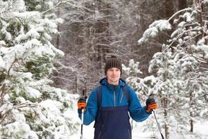 Skifahrer mit Rucksack und Hut mit Bommel mit Skistöcken in den Händen auf dem Hintergrund eines verschneiten Waldes. Langlaufen im Winterwald, Sport im Freien, gesunder Lebensstil, Wintersporttourismus. foto