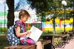 Mädchen mit Rucksack sitzt auf einer Bank und liest ein Buch in der Nähe der Schule. Schulanfang, Stundenplan, Tagebuch mit Noten. Bildung, Grundschulklassen, 1. September foto
