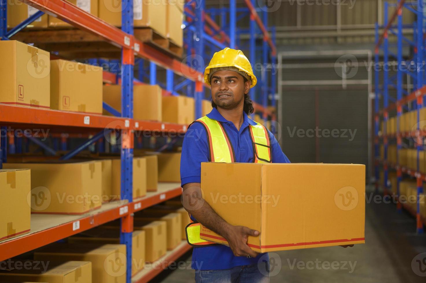 ung blandras manlig arbetare bär hjälm lyft kartong i lager, maskiner och logistik koncept. foto