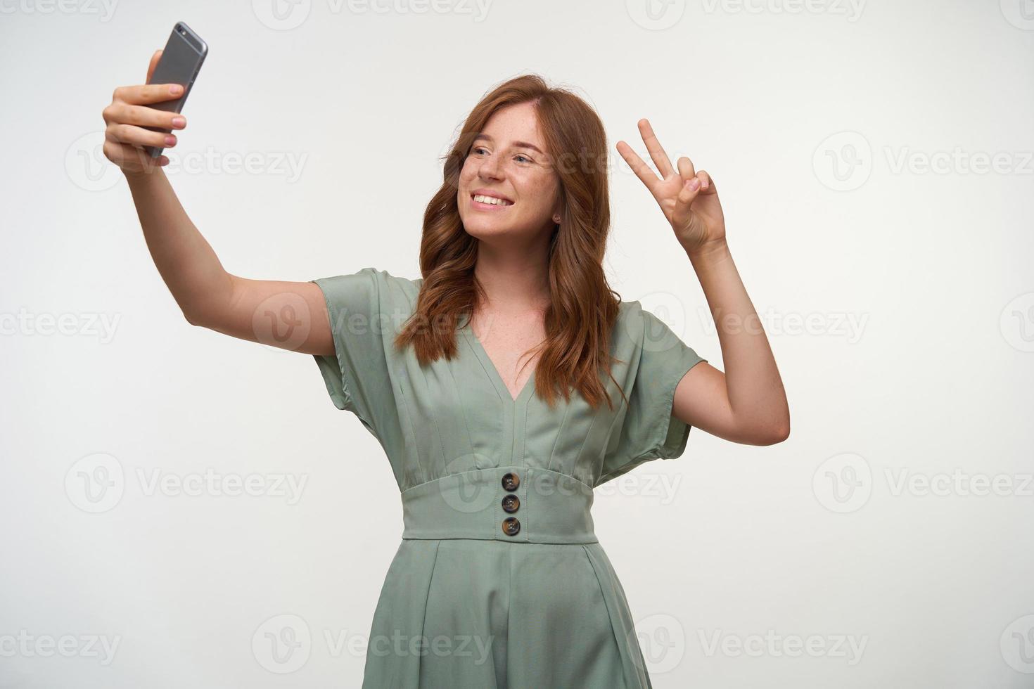 studiobild av charmig rödhårig ung kvinna som gör selfie med sin smartphone, ler glatt mot kameran och höjer två fingrar med fredsgest foto