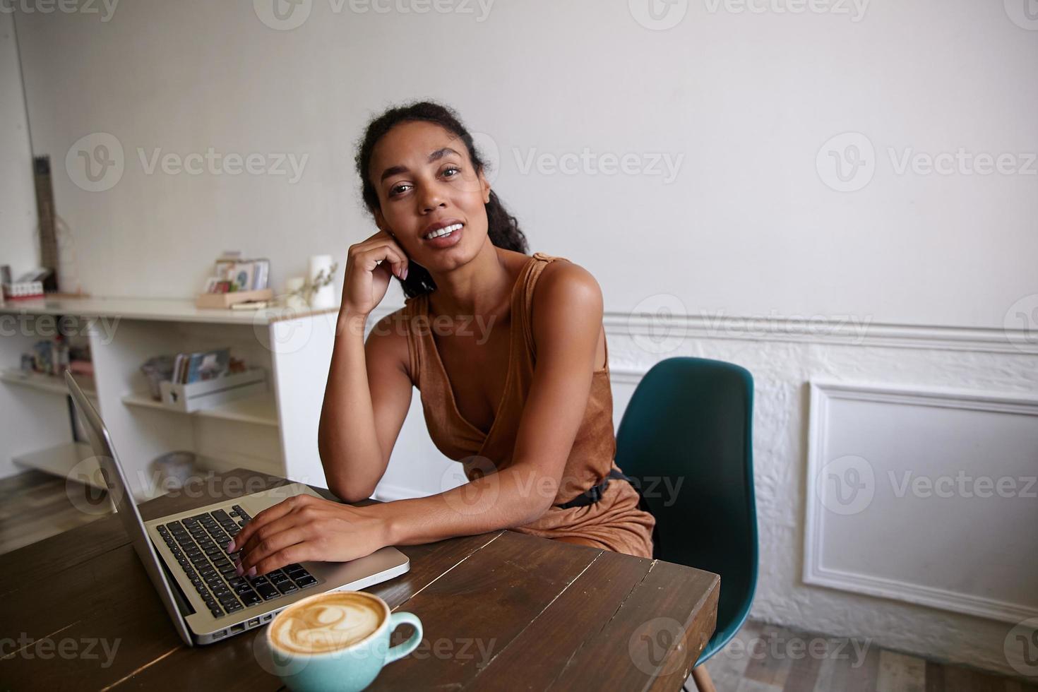 ung vacker mörkhyad kvinna poserar över kaféinteriören, dricker en kopp kaffe medan hon jobbar utanför kontoret, bär avslappnad klänning, är positiv foto