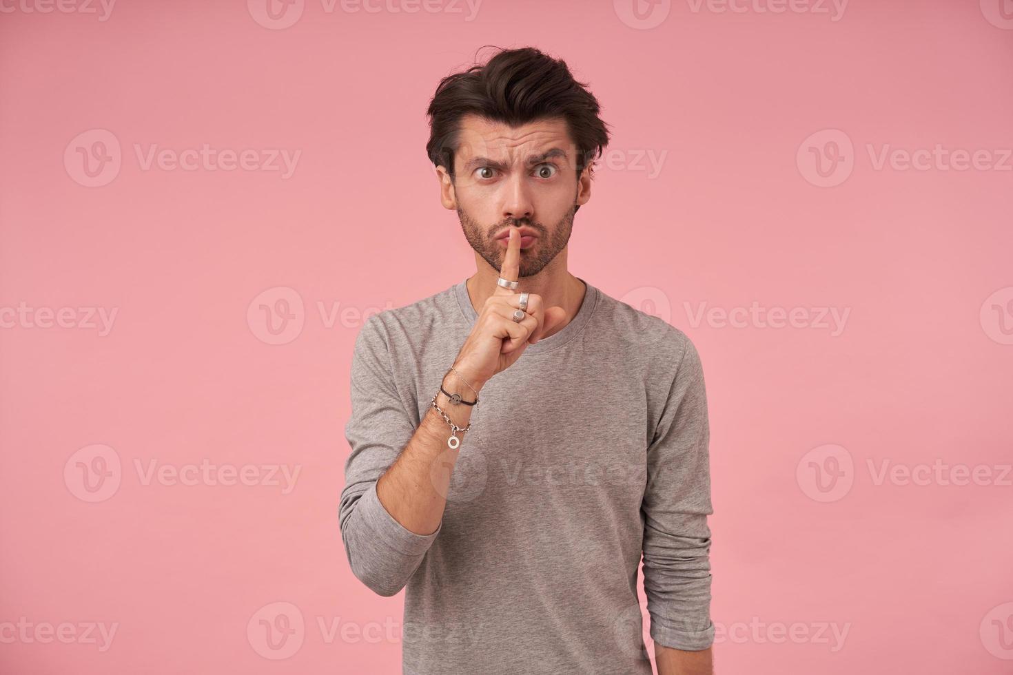 inomhusbild av ung attraktiv skäggig man i grå tröja som står över rosa bakgrund, höjer pekfingret till läpparna i tystnadsgest, ber att hålla hemlig foto