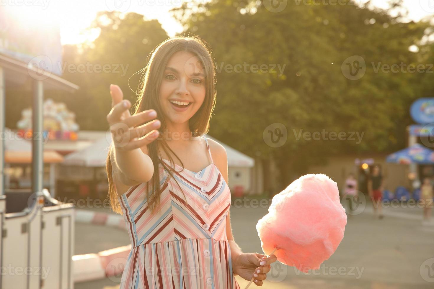 soligt utomhusfoto av charmig brunett med brunt hår som poserar över grön park med rosa sockervadd i handen, ger en hand och bjuder in att gå med henne och ler glatt mot kameran foto