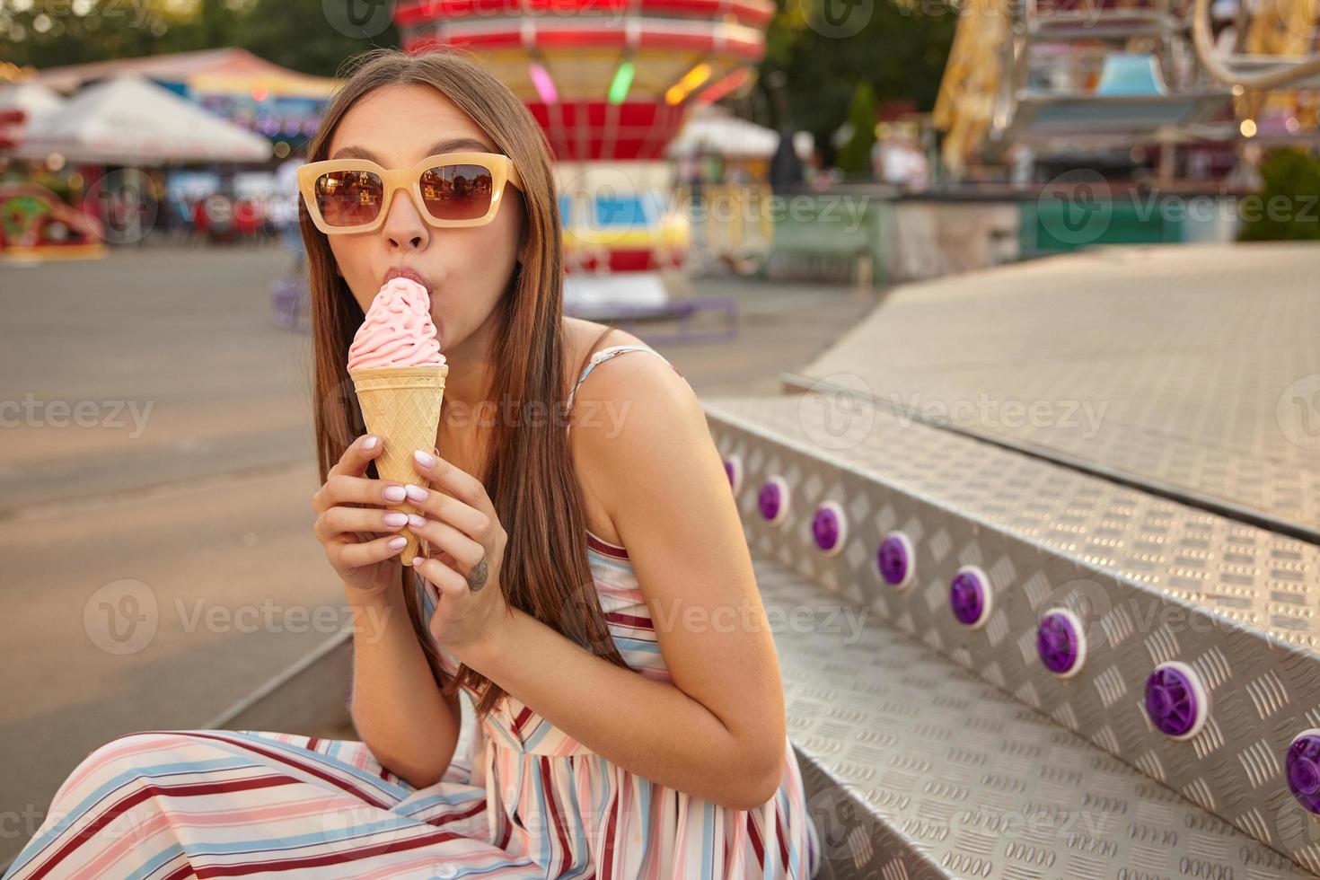 utomhusporträtt av en ganska ung långhårig brunettdam som sitter på trappan över nöjesparken, bär solglasögon och ljus sommarklänning, äter rosa glass i strut foto