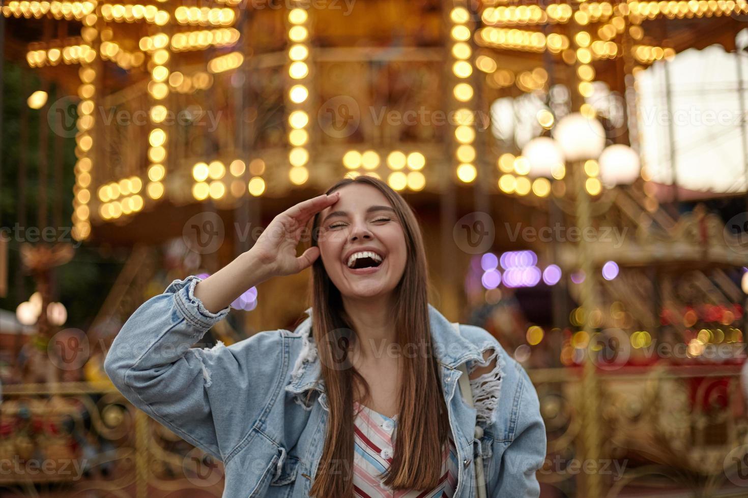 glad attraktiv långhårig kvinna i jeansrock poserar över karusellen i nöjesparken, ler full mun med slutna ögon och rör vid pannan med handen foto