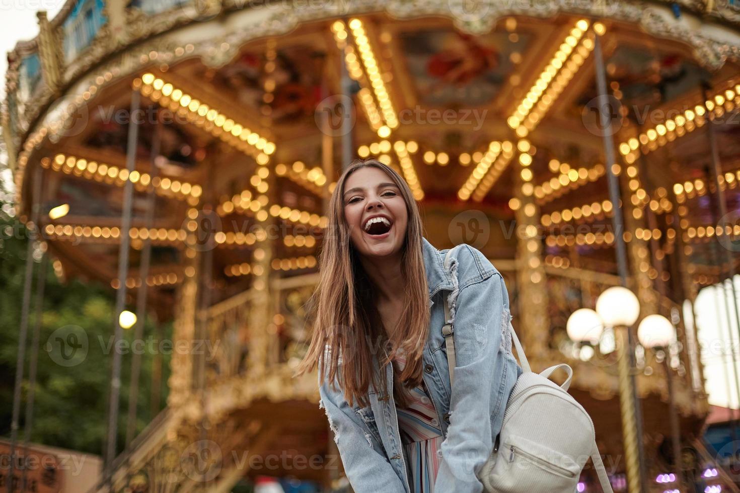 utomhusbild av glad ung vacker kvinna som bär jeansrock och vit ryggsäck, står över karusellen i nöjesparken och ler glatt mot kameran foto