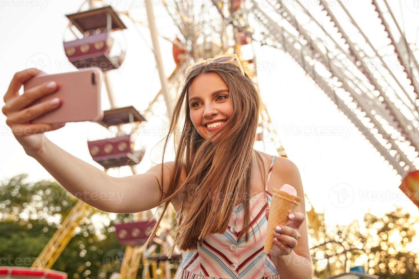 porträtt av glad ung härlig dam med charmigt leende som poserar över attraktioner i nöjespark, gör foto av sig själv med smartphone, håller en glassstrut i handen