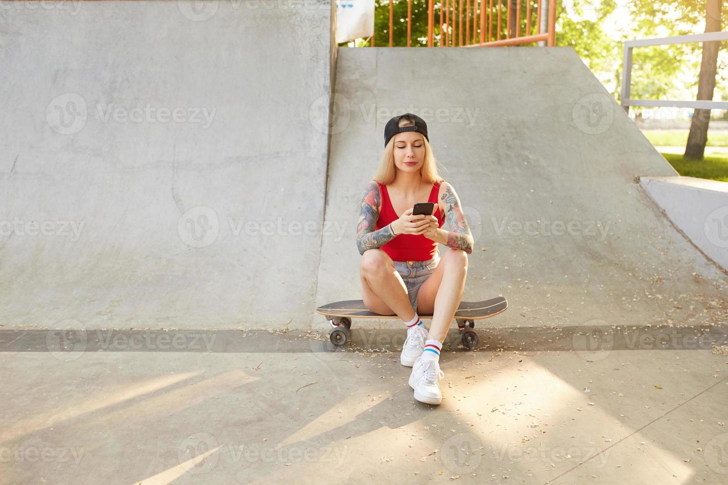utomhusfoto av vacker tatuerad ung blond kvinna i basebollkeps och vardagskläder, sitter över stadsparken en solig dag och tittar på skärmen på sin smartphone foto