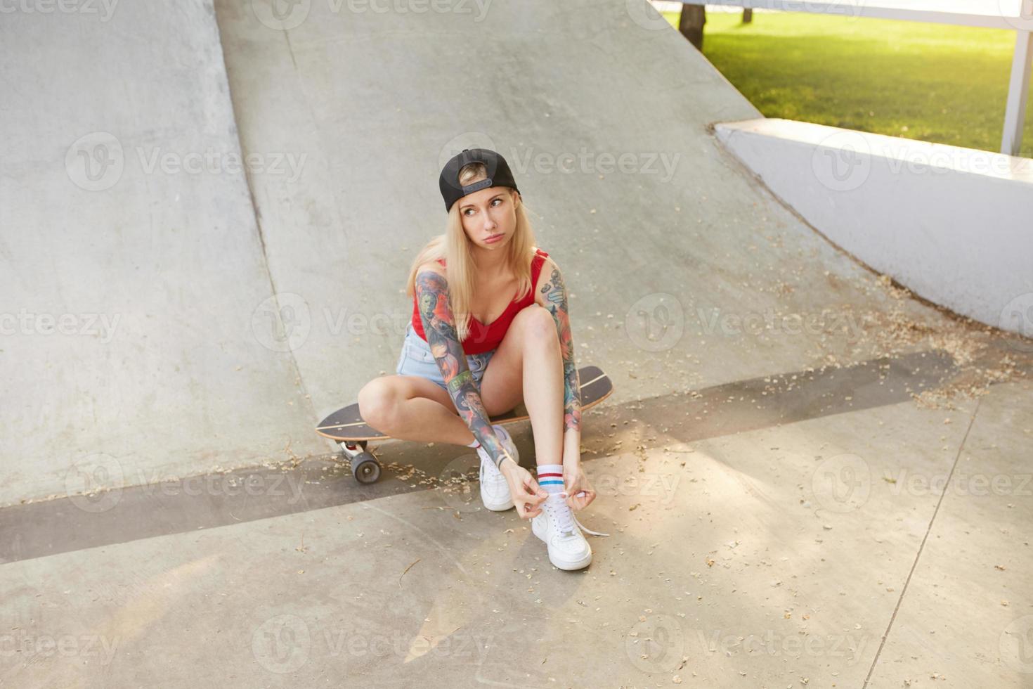 ung vacker blond kvinna med långt hår i basebollkeps och fritidskläder poserar utomhus en varm solig dag, knyter skosnören medan du sitter på skateboard foto
