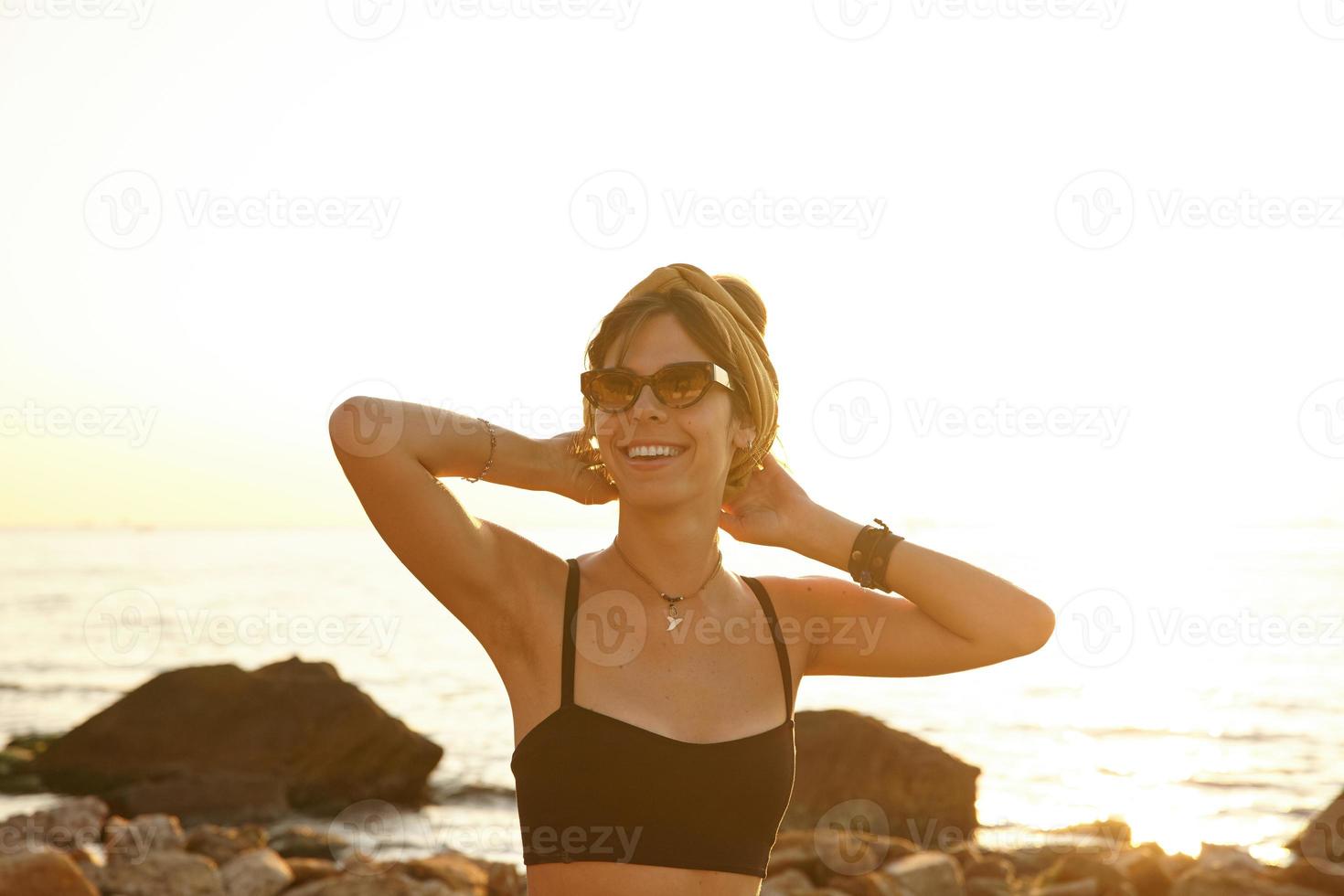 vacker ung mörkhårig kvinna med avslappnad frisyr poserar över strandpromenaden med höjda händer mot huvudet, tittar glatt mot kameran, bär svart topp och senapsfärgat pannband foto
