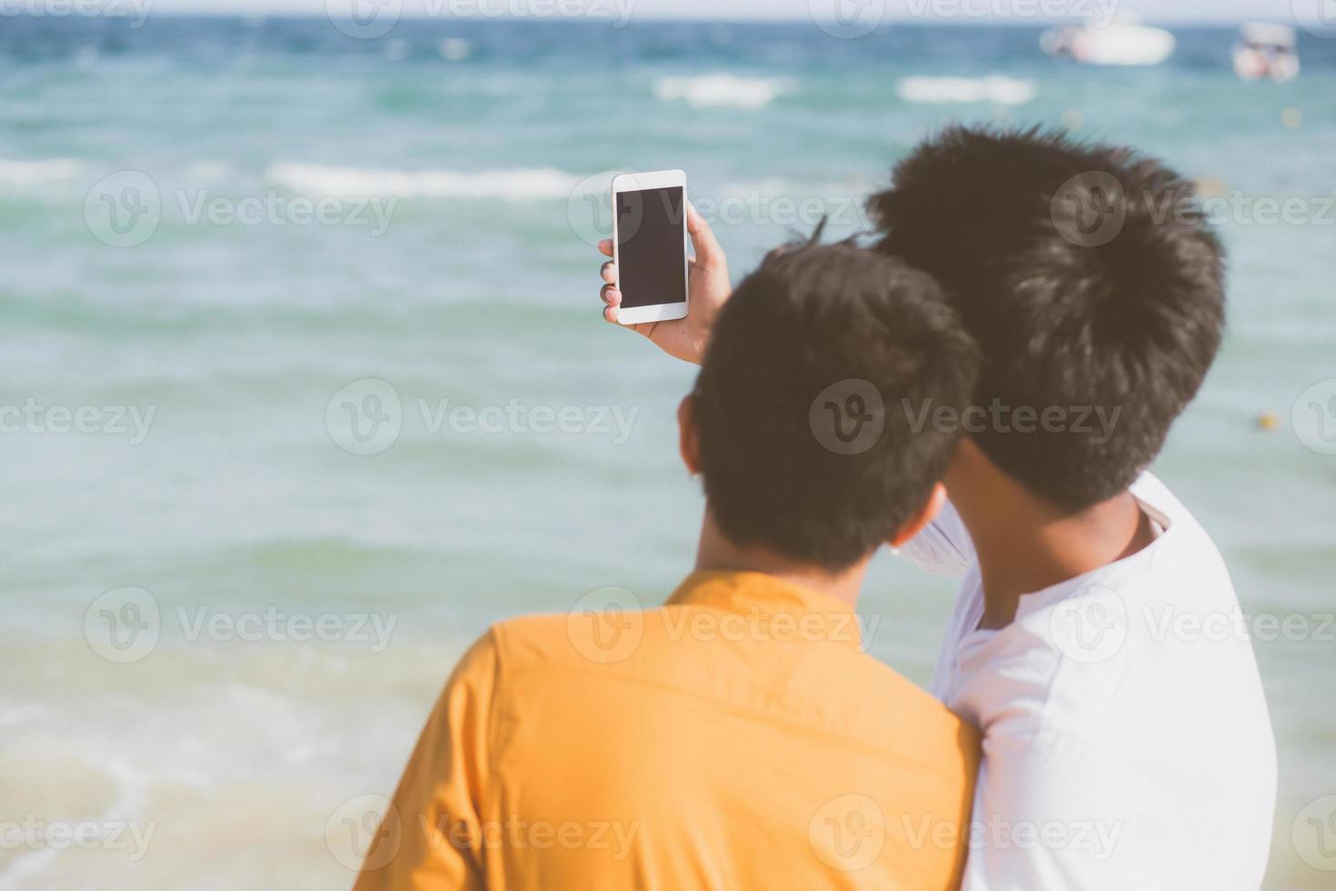 bakifrån gay porträtt ungt par leende tar en selfie foto tillsammans med smart mobiltelefon på stranden, lgbt homosexuell älskare i semestern till sjöss, två man kommer att resa, semester koncept.