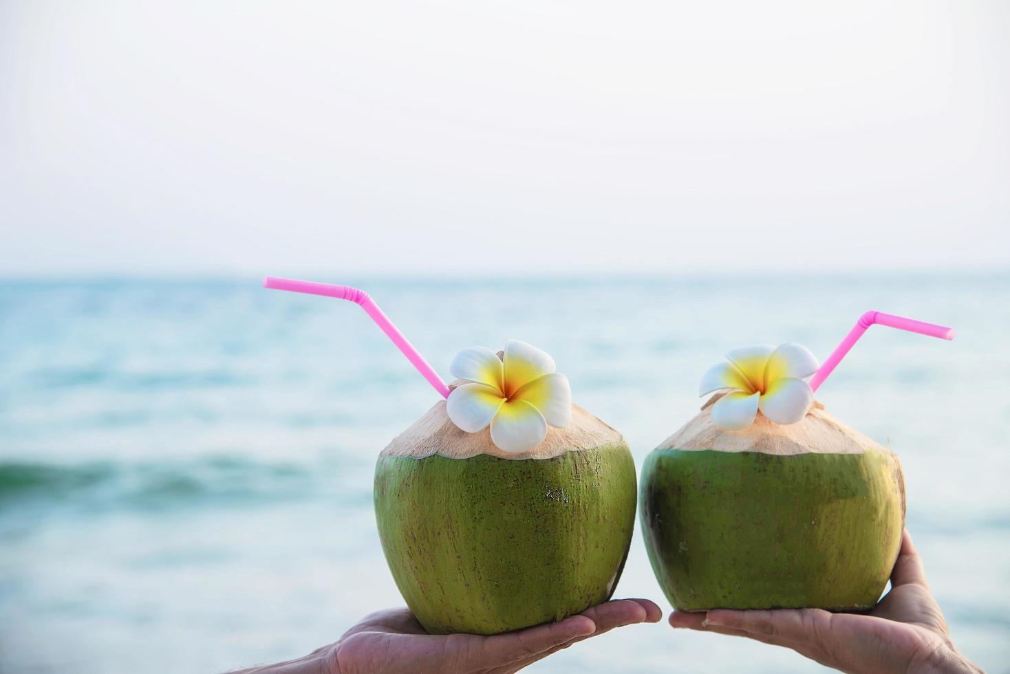 färsk kokosnöt i par händer med plumeria dekorerad på stranden med havsvågbakgrund - smekmånadsparturist med färsk frukt och havssand solsemesterbakgrundskoncept foto