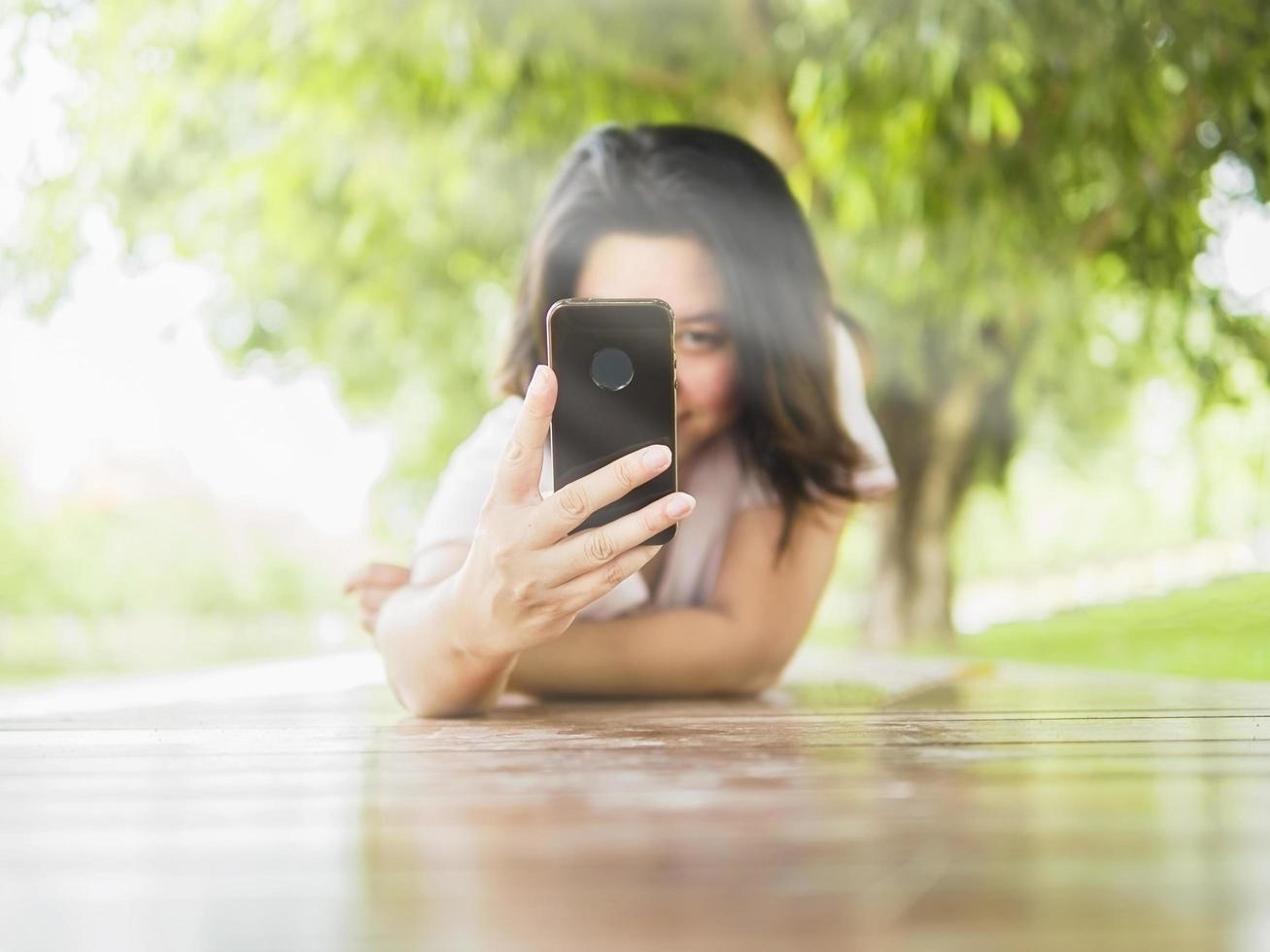 kvinna lägger sig på träterrass och tar foto med mobiltelefon i den gröna parken