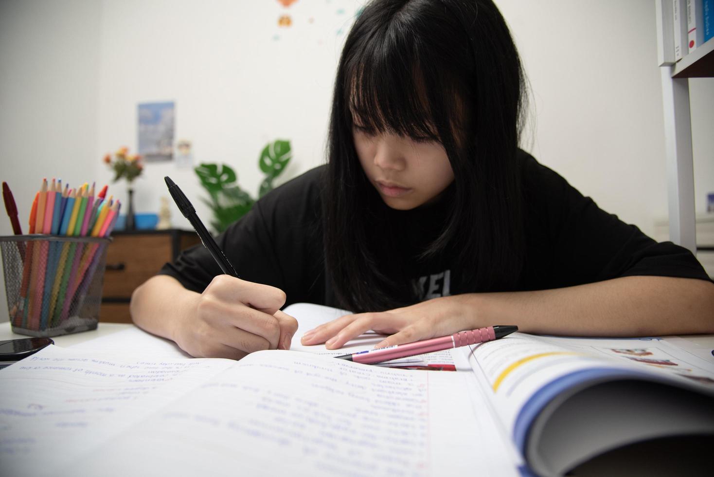 asiatisk studentflicka skriver läxor och läser bok vid skrivbordet foto