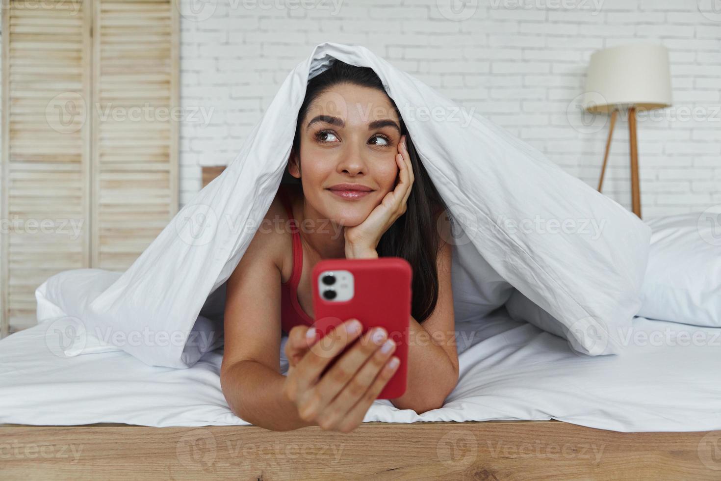 omtänksam kvinna tittar ut ur filten och håller smart telefon medan du ligger i sängen foto