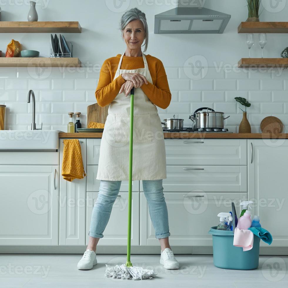 full längd av självsäker äldre kvinna som lutar sig mot moppen medan hon står vid det inhemska köket foto
