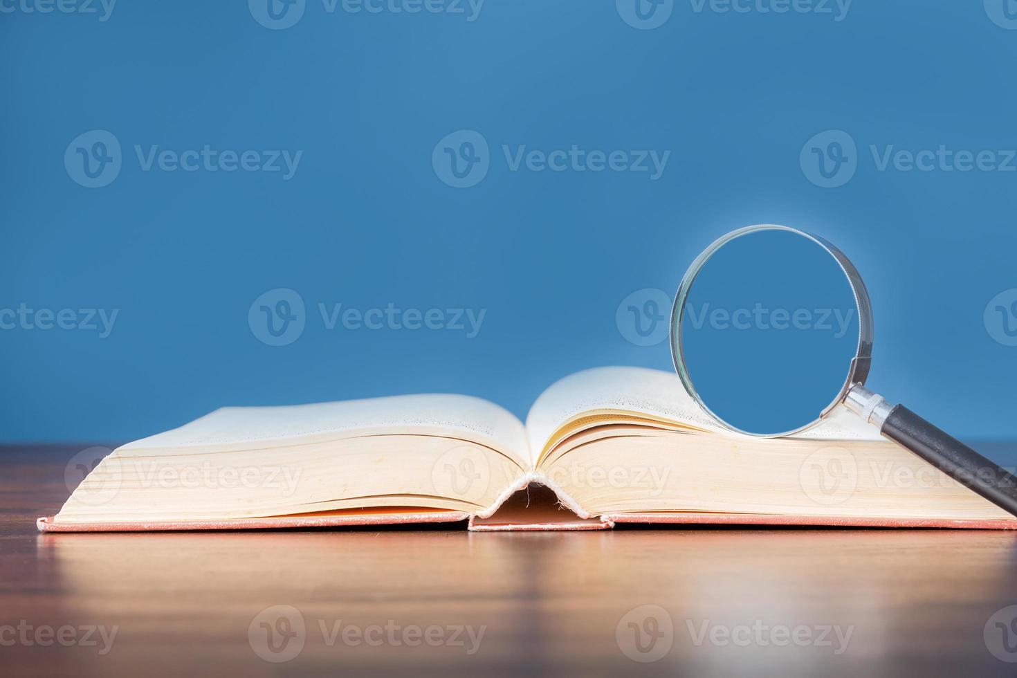 öppen bok med förstoringsglas på träskrivbord i informationsbibliotek för skolan eller universitetet, koncept för utbildning, läsning, studier, kopieringsutrymme och blå bakgrund. foto