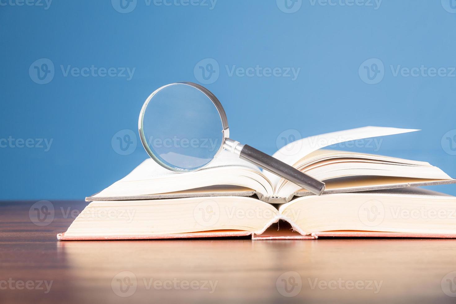 öppen bok med förstoringsglas på träskrivbord i informationsbibliotek för skolan eller universitetet, koncept för utbildning, läsning, studier, kopieringsutrymme och blå bakgrund. foto